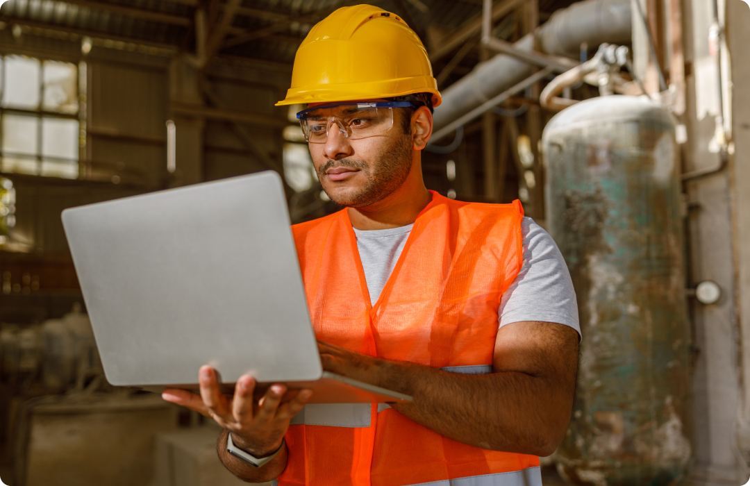Trabalhador em pé usando EPI - capacete amarelo, óculos de proteção e colete alaranjado - trabalhando com um computador nas mãos